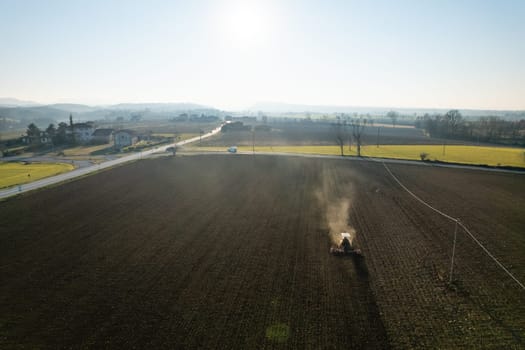 farmer driving a crawler tractor drags a cloud of dust behind, ploughing, power harrowing surface crust breaking to facilitate the birth and seeding of plants on dry soil