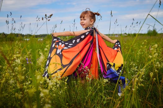 Portrait of little girl with Asian eyes and butterfly wings having fun and joy in meadow or field with grass, flowers on sunny summer day