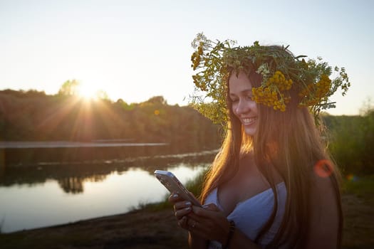 Young brunette girl in a white dress, sundress and wreath of flowers in summer on the coast of river or lake in the evening at sunset. Celebration of the Slavic pagan holiday of Ivan Kupala