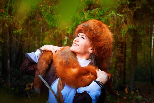 Girl in a leather jacket, a big red fox fur hat and with the skin of a fox killed on the hunt in the forest in autumn. A female model poses as fabulous royal huntress on nature hunt at photo shoot