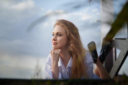 Beautiful girl with long hair in white shirt in open wooden pavillion in a village or small town. Young slender woman and sky with clouds on background on an autumn, spring or summer day