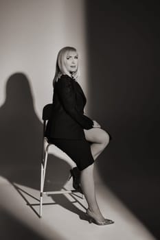 A fashionable woman in a black jacket and dress poses in a studio on a chair.