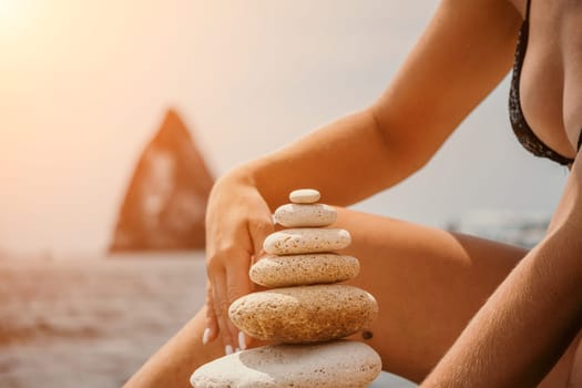 Woman bilds stones pyramid on the seashore on a sunny day on the blue sea background. Happy holidays. Pebble beach, calm sea, travel destination. Concept of happy vacation on the sea, meditation, spa