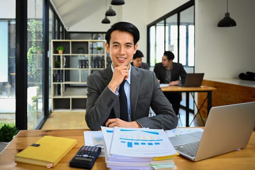 Male marketing business manager in formal clothes sitting at work desk with reports and laptop computer.