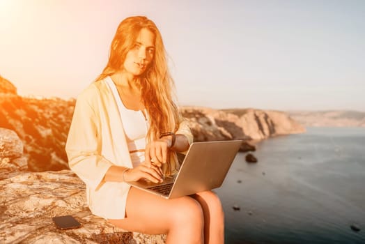 Successful business woman in yellow hat working on laptop by the sea. Pretty lady typing on computer at summer day outdoors. Freelance, travel and holidays concept.