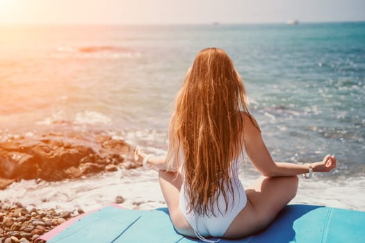 Young woman in swimsuit with long hair practicing stretching outdoors on yoga mat by the sea on a sunny day. Women's yoga fitness pilates routine. Healthy lifestyle, harmony and meditation concept.