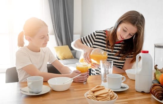 Girls sisters eat tasty breakfast together and filling glass with orange juice. Female child kid and subling with citrus beverage during morning meal