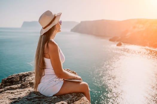 Successful business woman in yellow hat working on laptop by the sea. Pretty lady typing on computer at summer day outdoors. Freelance, travel and holidays concept.