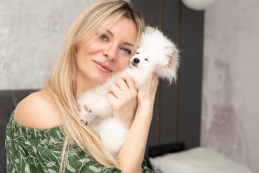 A beautiful young woman lies and plays on a bed in a home interior with a small domestic white and fluffy spitz dog. Concept. Close-up.