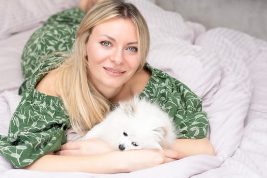 A beautiful young woman lies and plays on a bed in a home interior with a small domestic white and fluffy spitz dog. Concept. Close-up.