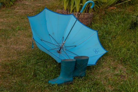 Blue rubber boots and a blue umbrella lies on the lawn. Protection from bad weather and rain in the garden. Rural life.