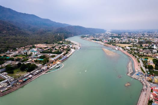 aerial drone shot of blue water of river ganga stretching into distance with himalayas with ram setu suspension bridge and temples on the banks of the river in India