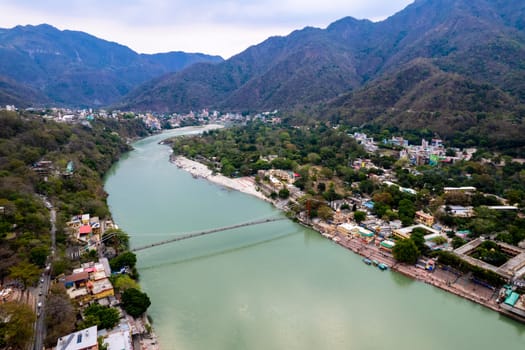 aerial drone shot of blue water of river ganga stretching into distance with himalayas with ram setu suspension bridge and temples on the banks of the river