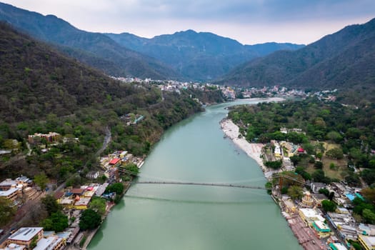 aerial drone shot of blue water of river ganga stretching into distance with himalayas with ram setu suspension bridge and temples on the banks of the river