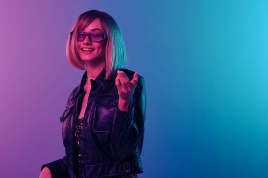 Cute cheerful blonde woman in leather jacket sparkly dress trendy sunglasses point hand at camera look aside posing isolated in blue pink color light studio background. Neon party concept. Copy space