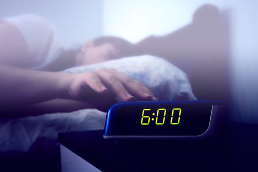 A sleeping woman's hand reaches for a digital alarm clock on the bedside table. Color toning and haze filter on the image