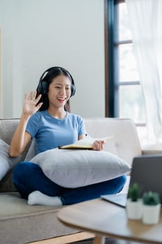 Asian young business woman is sitting on the sofa is wearing headphones video call conference with laptop computer raise hand saying hi team at home office, work from home