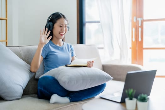 Asian young business woman is sitting on the sofa is wearing headphones video call conference with laptop computer raise hand saying hi team at home office, work from home