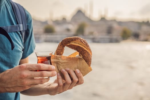 Man in Istanbul having breakfast with Simit and a glass of Turkish tea. Glass of Turkish tea and bagel Simit against golden horn bay and the Galata Tower in Istanbul, Turkey. Turkiye.