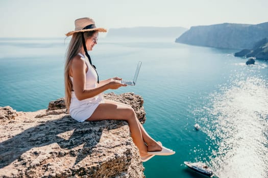 Successful business woman in yellow hat working on laptop by the sea. Pretty lady typing on computer at summer day outdoors. Freelance, travel and holidays concept.