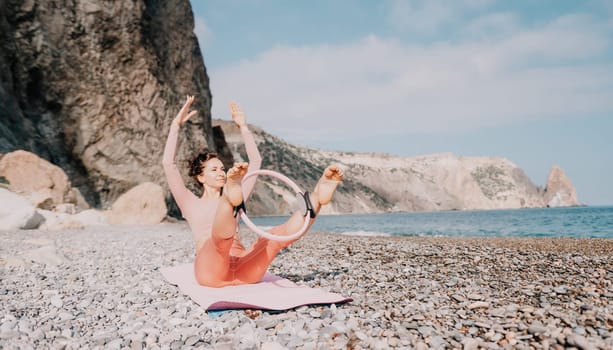 Middle aged well looking woman with black hair doing Pilates with the ring on the yoga mat near the sea on the pebble beach. Female fitness yoga concept. Healthy lifestyle, harmony and meditation.