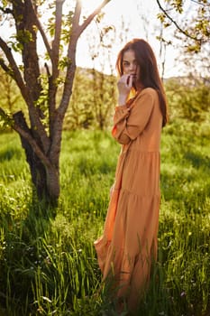 a sweet, thoughtful woman stands in nature near a tree in a long orange dress, illuminated from the back by the sunset rays of the sun and holding her hand near her face looks away. High quality photo