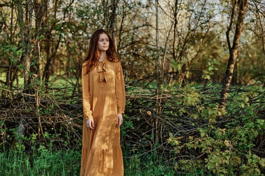 a beautiful woman walks in the shade near the trees, dressed in a long orange dress, enjoying the weather and the weekend. The theme of privacy with nature, horizontal photography on the street. High quality photo