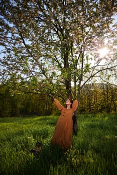 a slender, attractive woman with long red hair stands in the countryside near a flowering tree in a long orange dress and holding a branch with her hands loosely leans to the side. Vertical photography. High quality photo