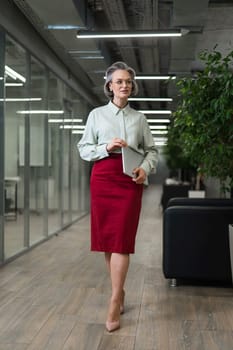 Attractive mature caucasian woman holding laptop while standing in office