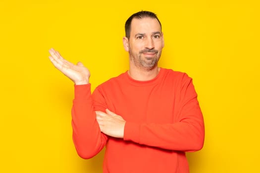 Hispanic man with a beard and red sweater in a friendly attitude offering something on the palm of his hand, isolated on a yellow background.