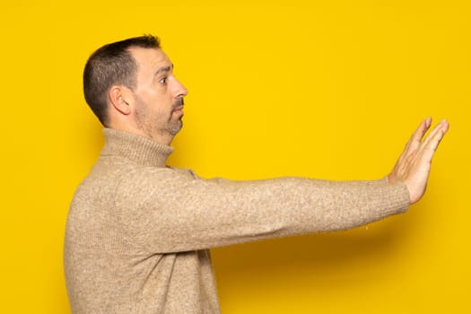 Side view of a scared man standing with a scared or worried face, looking at the camera and blocking with his hands. Indoor studio shot isolated on yellow background.