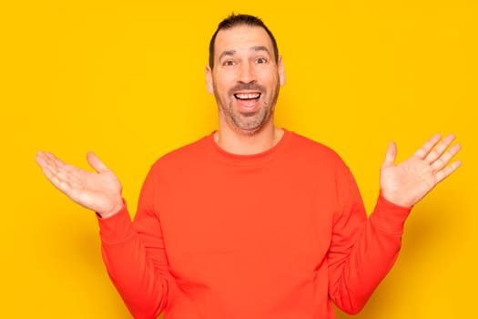 Funny smiling man in red sweater showing palms up, unexpected surprise gesture isolated over yellow background. Attractive happy man listening to good news, catching something