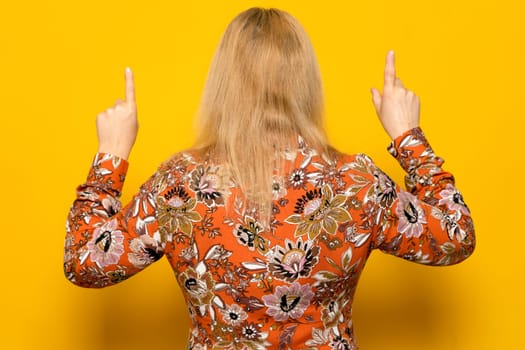 Caucasian woman in casual sweater posing backwards pointing up with index fingers turned away from the camera, isolated over yellow background