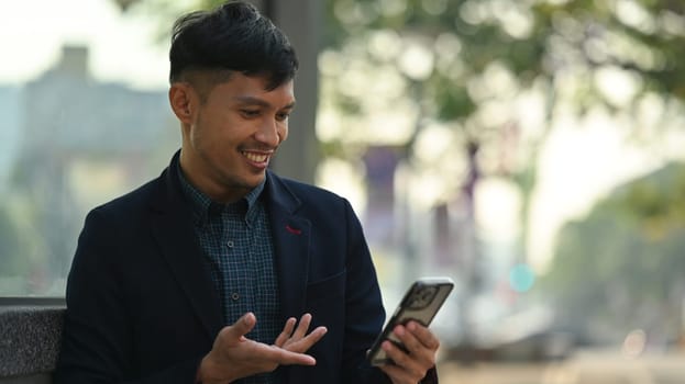 Positive businessman or worker dressed in formal clothes sitting outdoors in urban modern city and using smartphone.