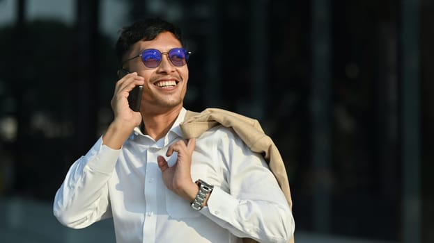 Smiling young businessman standing outside building with jacket over his shoulder and talking on mobile phone.