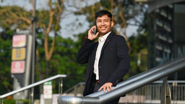 Handsome businessman talking on mobile phone while standing outdoor with cityscape background in early morning.