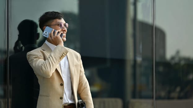 Portrait of adult man in elegant business suit having phone conversation while standing in front of modern office building.