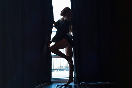 Striptease at home. Sexy blonde undressing at the window. Silhouette of a seductive girl.