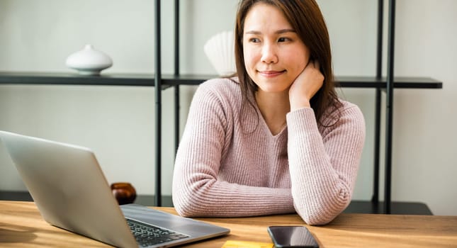 Dreamy businesswoman sitting at work desk in office thinking of new project strategy or vacation, Asian business woman making serious decision she work on laptop computer
