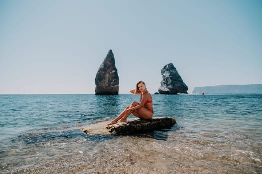 Woman travel sea. Young Happy woman in a long red dress posing on a beach near the sea on background of volcanic rocks, like in Iceland, sharing travel adventure journey