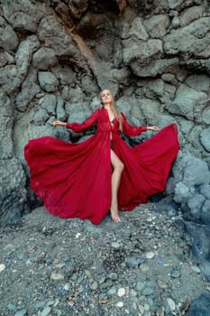 Red dress rocks woman. A blonde with flowing hair in a long flowing red dress stands near a rock of volcanic origin. Travel concept, photo session at sea.