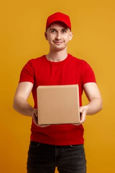 Delivery man employee in red cap, red t-shirt holding cardboard box on yellow background. focus on box