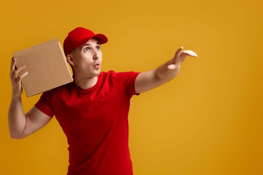Delivery man employee in red cap, red t-shirt holding cardboard box and pointing finger at the side isolated on yellow background