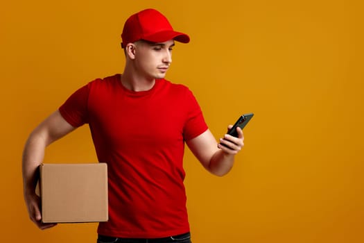 Delivery man employee in red cap, empty t-shirt holding empty cardboard box and phone isolated on orange background