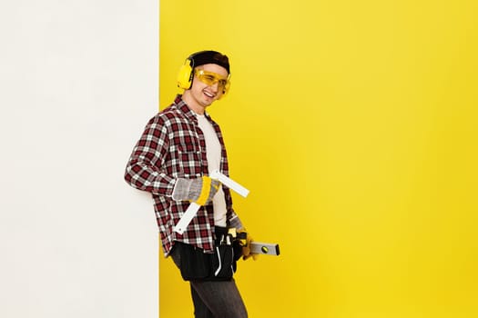winking worker man or builder in construction helmet near white banner on yellow background. building carpentry profession