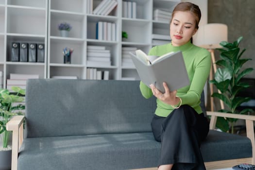 Beautiful young female for early in morning, writes notes in notepad, enjoys hot coffee from paper cup, sits on comfortable sofa, cozy domestic atmosphere