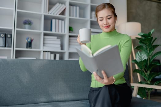 Beautiful young female for early in morning, writes notes in notepad, enjoys hot coffee from paper cup, sits on comfortable sofa, cozy domestic atmosphere