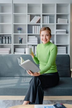 Beautiful young female for early in morning, writes notes in notepad, enjoys hot coffee from paper cup, sits on comfortable sofa, cozy domestic atmosphere