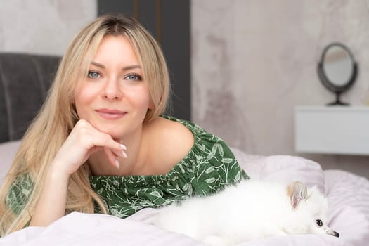 A beautiful young woman lies and plays on a bed in a home interior with a small domestic white and fluffy spitz dog. Concept. Close-up.