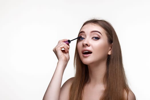 Beautiful young woman applying black mascara on long thick eyelashes with a brush and making night makeup on white background